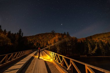 Dağ nehri üzerindeki köprüde, yıldızlı gökyüzünün altında fenerli bir kadın. Yüksek ISO 'dan bazı sesler var