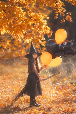 Süpürgeli, siyah turuncu balonlu, uzun kahverengi saçlı güzel bir kız sonbahar ormanında yürüyor.