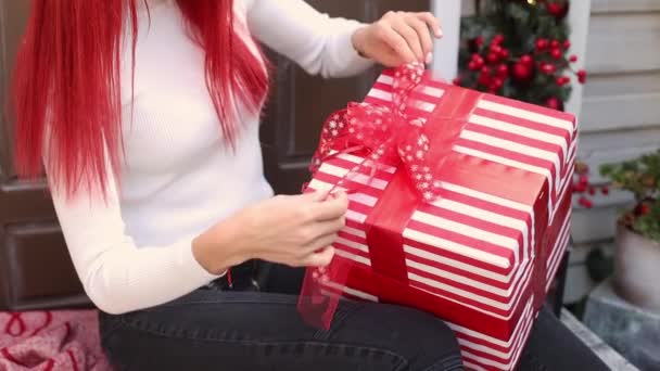 Zbliżenie hipsterskie dziewczyny ręce z długimi, jasnoczerwonymi włosami przygotowujące się na zimowe wakacje i pakujące prezent świąteczny z kokardą na werandzie domu ozdobionego wieńcem i girlandą — Wideo stockowe