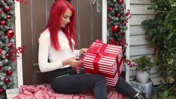 Portrait of a hipster young woman 30 years old with bright red hair preparing for winter holidays and packing Christmas present on the porch of the house decorated with a wreath and garland — Stock Video