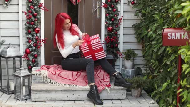 Koko kehon muotokuva nuoresta 30-vuotiaasta naisesta, jolla on kirkkaan punaiset hiukset, jotka valmistautuvat talvilomiin ja pakkaavat joululahjan talon kuistilla, joka on koristeltu seppeleellä ja seppeleellä. — kuvapankkivideo