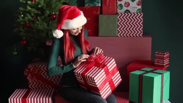 冬の休日のために準備し 弓でクリスマスプレゼントを詰め 多くの贈り物の中で装飾されたクリスマスツリーの下に座っている明るい赤い髪で30歳のフリーク若い女性の肖像画 — ストック動画