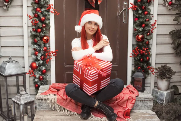 Retrato de cuerpo completo de mujer joven de 30 años con pelo rojo brillante en sombrero de santa preparación para las vacaciones de invierno, embalaje regalo de Navidad en el porche de la casa decorada con una corona y guirnalda —  Fotos de Stock