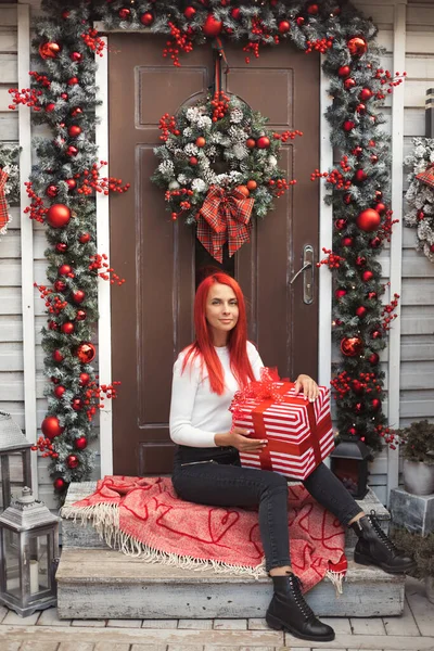 Ritratto completo di una giovane donna di 30 anni con i capelli rossi brillanti che si prepara per le vacanze invernali, imballando il regalo di Natale sul portico della casa decorato con ghirlanda e ghirlanda. — Foto Stock