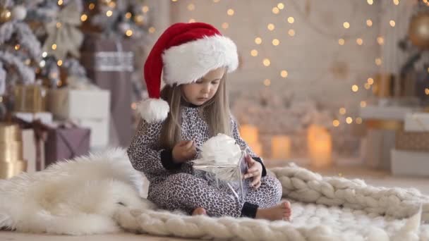 Niña linda en pijama y sombrero de santa en casa abre su regalo de Navidad — Vídeos de Stock
