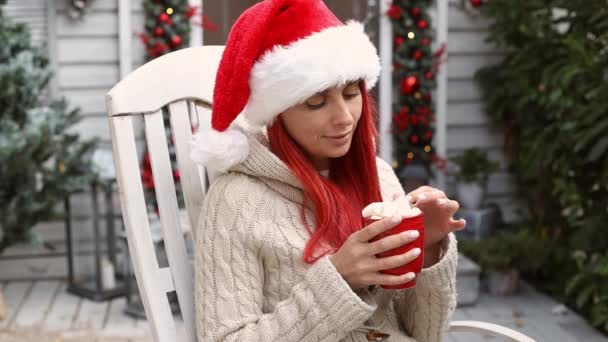 Gyönyörű hipszter lány piros hajjal, télapó sapkában ül egy hintaszékben a ház verandáján, élvezi a téli ünnepeket és karácsonyi hangulatot, és kakaót iszik mályvacukorral. 