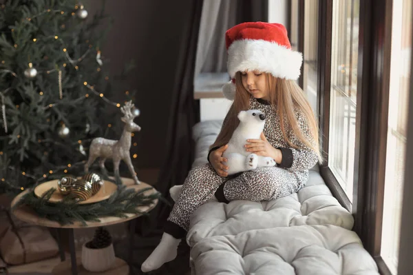 Pełna długość cute little girl 3-4 lat w Santa Claus kapelusz i piżama siedzi w domu na parapecie przy oknie w pięknym szarym wnętrzu ozdobione choinką w tle — Zdjęcie stockowe