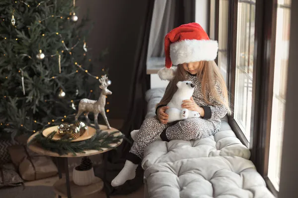 Gezichtsloos portret van schattig klein meisje 3-4 jaar oud in kerstman hoed en pyjama zit thuis op de vensterbank door het raam in mooie grijze interieur versierd met een kerstboom op de achtergrond — Stockfoto