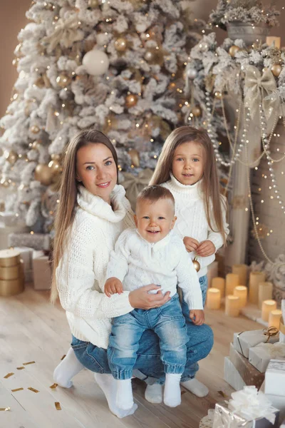 Krásná bělošská rodina: matka a dvě děti malý syn 1 rok a malá dcera 3-4 roky, slaví Vánoce doma a objímají spolu v útulných bílých svetrech a džínách. — Stock fotografie