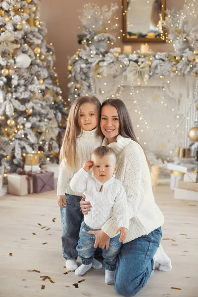 Güzel bir beyaz aile: anne ve iki çocuk 1 yaşında küçük oğlan ve 3-4 yaşlarında küçük bir kız, Noel 'i evde kutluyorlar ve rahat beyaz kazaklar ve kot pantolonlar içinde birbirlerine sarılıyorlar..