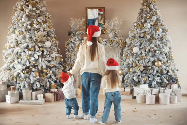 Krásná rodina zezadu: matka a dvě děti, oslavit vánoční den doma spolu v útulné bílé svetry, džíny a Santa Claus klobouky. — Stock fotografie