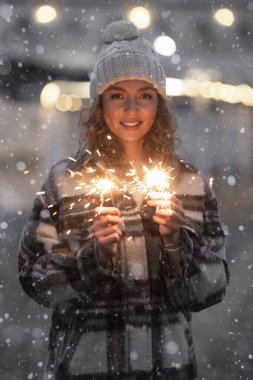Çekici ve gülümseyen beyaz kadın portresi. 25-30 yaşlarında, moda modasına uygun ceketli ve örgü şapkalı. Yeni yıl partisini sokakta havai fişekli bengal ışıklarıyla kutluyor.