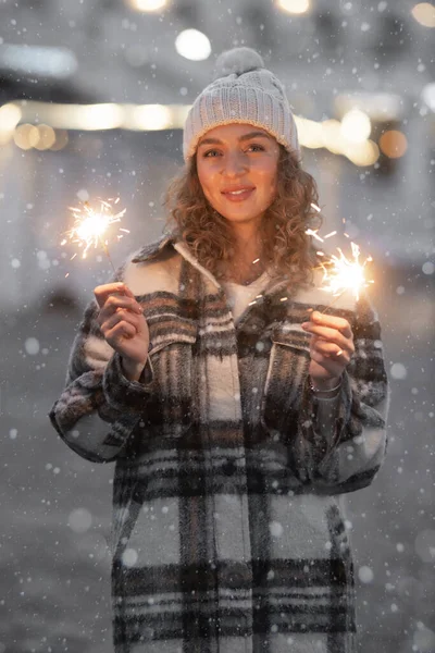 Ritratto verticale di bella donna riccia 25-30 anni che tiene in mano una scintilla ardente su neve città notte ligths sfondo Immagine Stock