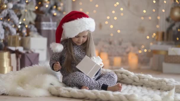 Une petite fille mignonne de 3-4 ans en pyjama et chapeau de Père Noël se trouve à la maison sous un arbre de Noël et ouvre son cadeau de vacances sur le fond de lumière de la cheminée — Video