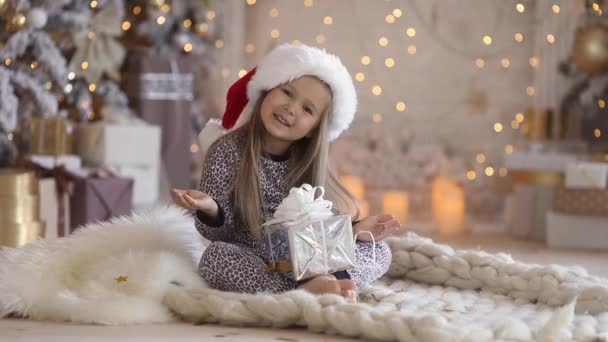 Une petite fille mignonne de 3-4 ans en pyjama et chapeau de Père Noël se trouve à la maison sous un arbre de Noël et ouvre son cadeau de vacances sur le fond de lumière de la cheminée — Video