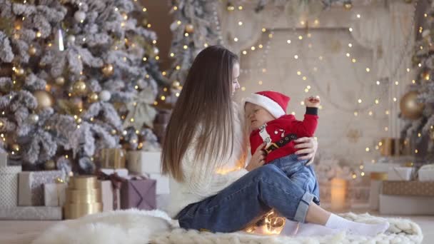 漂亮的年轻家庭：30岁至35岁的女人，穿着休闲舒适的白色毛衣、牛仔裤和圣诞老人帽，坐在圣诞树下，和她一岁的儿子一起玩着滑稽的圣诞老人服装 — 图库视频影像
