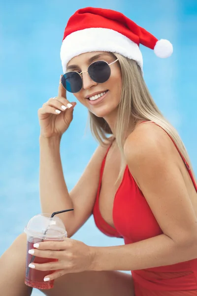 Mulher loira feliz bonita 25-30 anos de idade em um maiô vermelho e óculos de sol está descansando no hotel no resort à beira da piscina. Menina feliz se divertir na festa de Natal com coquetel em chapéu de santa claus. — Fotografia de Stock