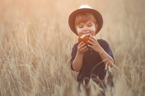 Cute and funny little boy 2-3 years old in a straw hat eating a fresh sweet bun in a wheat field on a summer day at sunset — Stock Photo, Image