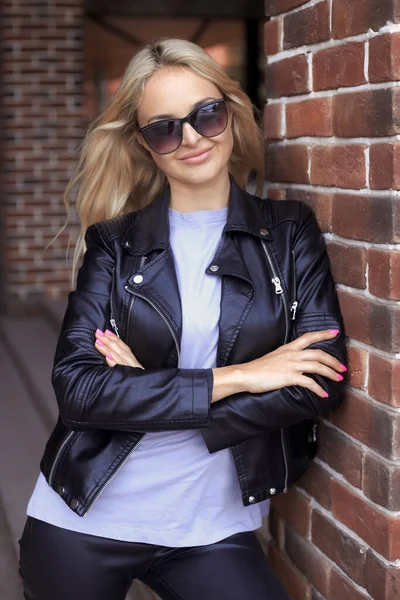 Vertical urban portrait of stylish beautiful millennial woman 25-30 years old with long hair in sunglasses, black leather jacket and purple t-shirt standing near the brick wall in the city — Stock fotografie