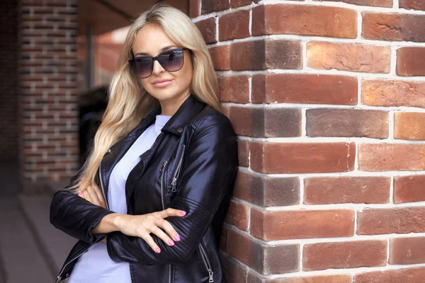 Urban portrait of stylish beautiful caucasian millennial woman 25-30 years old with long hair in sunglasses, black leather jacket and purple t-shirt standing near the brick wall in the city — Stockfoto