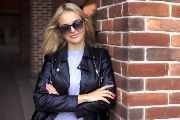 Urban positive portrait of stylish beautiful caucasian millennial woman 25-30 years old with long hair in sunglasses, black leather jacket and purple t-shirt standing near the brick wall in the city — Stock fotografie