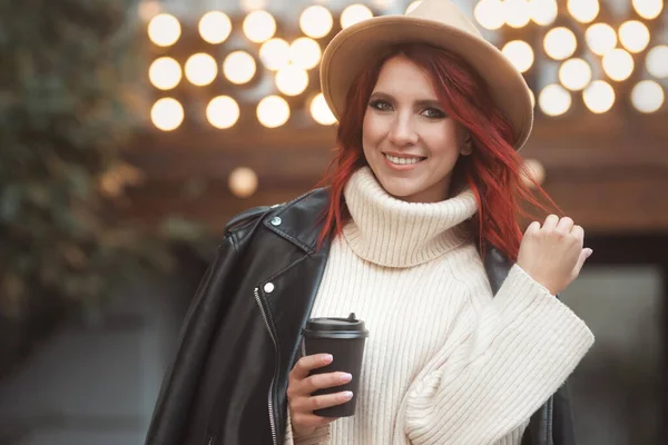 25-30 yaşlarında şık ve şık kızıl saçlı bir kadının sokak tarzı portresi. Günlük moda kıyafetleri içinde, açık kafede oturuyor ve kağıt bardaktan sıcak kahve içiyor.