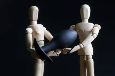 Two wooden figurines, anthropomorphic mannequins, hold a black silicone butt plug. Element of an intimate bedroom interior. Sex toys concept. Close-up