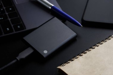 Bilgisayarın, akıllı telefonun, not defterinin ve kalemin yanındaki siyah arkaplandaki harici taşınabilir SSD diski. Sağlam bir State Drive. Modern teknolojiler, cihazlar ve SSD disk kullanımı kavramı. Yakın plan.