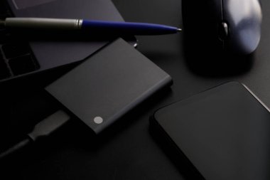Bilgisayar, kalem ve bilgisayar faresinin yanındaki siyah arka planda akıllı telefon ve harici taşınabilir SSD. Sağlam bir State Drive. Modern cihazlar ve bilgi depolama kavramı. Yakın plan.