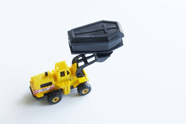 Yellow toy forklift is holding a black toy coffin in a bucket. Concept of the exhumation coffin and forensic examination. Black humor. Free space for an inscription. Selective focusing. Close-up