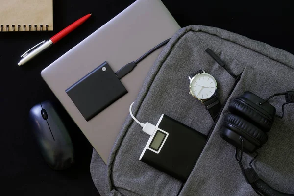 Laptop, harici SSD, güç bankası, kablosuz kulaklık, bilgisayar faresi ve kol saati olan modaya uygun tekstil şehir sırt çantası. Siyah arka plan. Seyahat, seyahat ve aletlerle yaşam tarzı. Yakın plan.