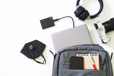 Laptop, akıllı telefon, aynasız kamera, kulaklıklar, harici SSD, koruyucu tıbbi solunum cihazı ve gri sırt çantası. Coronavirüs salgınında seyahat, gezi ve çalışma araçları kavramı. Yakın plan.