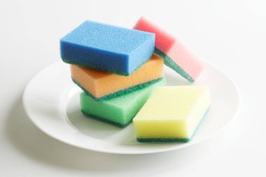 Beş tane çok renkli bulaşık süngeri ve açık arkaplanda beyaz bir tabak. Bulaşık yıkamak ve mutfağı temizlemek. Gün ışığı