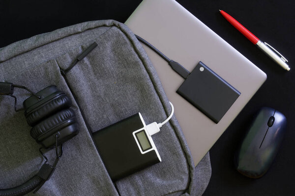 Серый рюкзак с ноутбуком или ультрабуком, внешний SSD и банк питания, беспроводная мышь и наушники. Путешествия, поездки и образ жизни с гаджетами. Чёрный фон. Крупный план