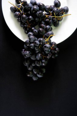 Siyah arka planda beyaz bir tabakta nem damlacıklarıyla kaplı siyah üzümler. Yakın plan.