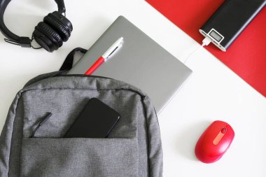 Laptop, akıllı telefon, elektrik bankası, cep telefonu, kulaklık, fare, kalem ve gri sırt çantası ya da çanta. Beyaz ve kırmızı arka plan. Seyahat, seyahat, eğitim ve aletlerle yaşam tarzı. Cihazlar şarj ediliyor. Yakın plan.