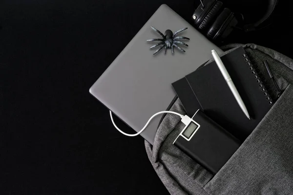 銀色のラップトップは 暗いテーブルの上に灰色のバックパックにあります 近くにはパワーバンク ノートパソコン 噴水ペン ヘッドフォン おもちゃのクモがあります 生活をガジェットで — ストック写真