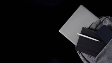 Silver laptop, along with smartphone, notepad and fountain pen, lies in a gray textile backpack on a dark table. Travel, trip, study and lifestyle with gadgets. Copy space. Close-up
