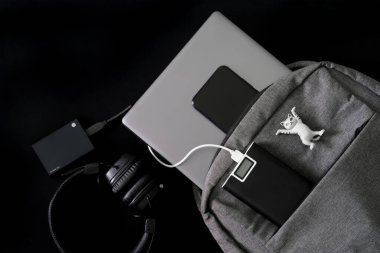 Silver laptop in a gray backpack on a dark table. Nearby are power bank, headphones, smartphone, external portable ssd and toy mascot cat. Travel, trip, study and lifestyle with gadgets. Close-up