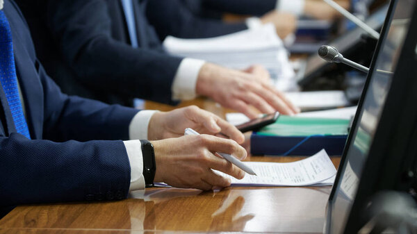 Hands of men - businessmen, politicians, managers or lawyers, sitting at table with paper documents, microphones and monitors. Concept of teamwork, negotiations or conference. Selective focus. No face