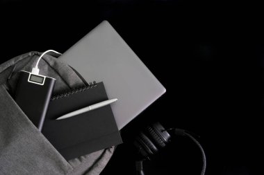 Gümüş rengi dizüstü bilgisayar gri sırt çantasında duruyor. Yanında not defteri, kalem ve harici batarya var. Güç bankası, kulaklıkların yanında. Siyah arka plan. Yazı için uzayı kopyala. Seyahat et ve aletlerle çalış. Yakın plan.