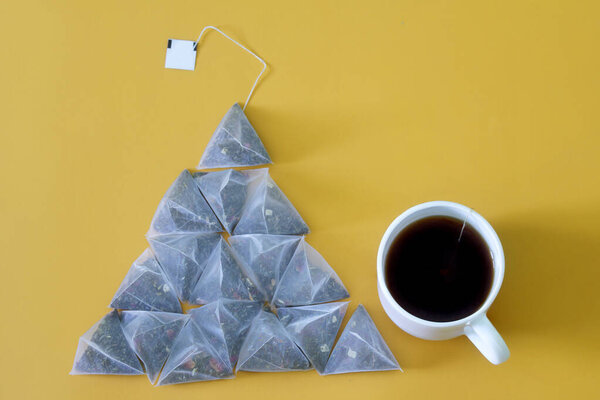Lot of pyramid tea bags with black and green tea, with pieces of fruit, and a white mug on a yellow background. Pyramid shape. Healthy food and drink concept. Close-up