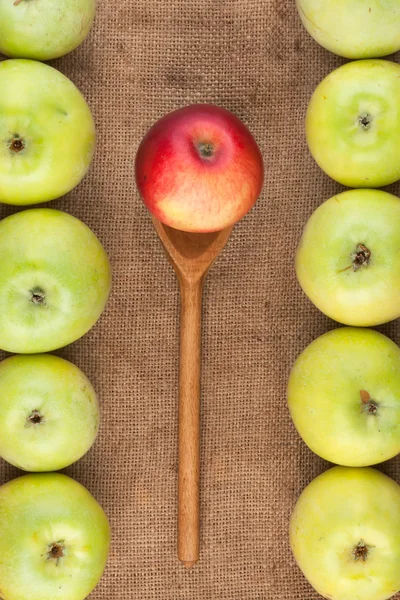 Spoon with red apple lying on the sackcloth among green apples — Stock Photo, Image