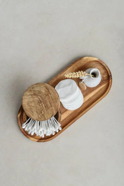 Cotton buds in wooden box with round facial cotton pads on wooden tray on a light concrete background, top view