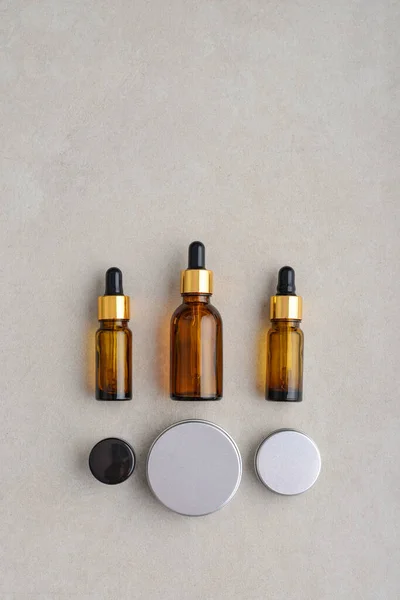 Dropper brown glass bottles, metal round tin cosmetic cans and small plastic can on light concrete background, top view