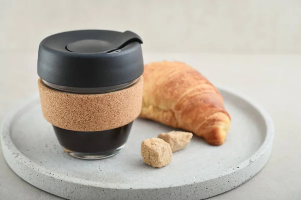 Coffee to-go in reusable travel mug made from glass and cork band with croissant on tray. Zero waste. Sustainable lifestyle concept.