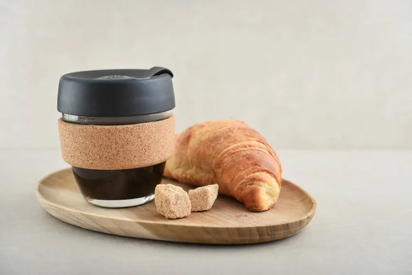 Coffee to-go in reusable travel mug made from glass and cork band with croissant on tray. Zero waste. Sustainable lifestyle concept.