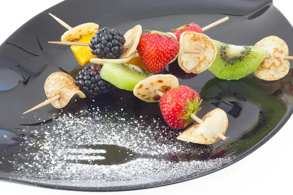 Mini blini (crêpe) aux fruits coupés sur brochettes, sur assiette noire — Photo