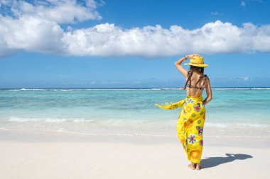 Sea Beach 'te mavi gökyüzüne bakan genç bir kız, sarı kanat çırpan elbiseli bir kız, arka plan manzaralı, yaz tatili konsepti. 