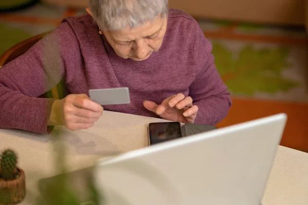 Gri saçlı son sınıf öğrencisi bir kadın elinde kredi kartıyla dizüstü bilgisayarında oturuyor ve internetten alışveriş yapıyor. Çevrimiçi alışveriş kavramı